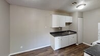 This single-floor residential unit offers a practical and well-organized layout designed for comfortable everyday living. The living area is finished with plush carpet underfoot and neutral-toned walls, creating a warm and inviting atmosphere. A window fitted with vertical blinds allows natural light to filter into the space, enhancing the open feel of the room. The layout flows naturally from the living area toward the kitchen and hallway, with transitions between carpet and dark hardwood-style flooring that add visual contrast throughout.

The galley-style kitchen is efficiently designed, featuring white cabinetry with brushed hardware, dark granite-look countertops, a stainless steel sink, and a full suite of white appliances including a refrigerator, electric range, and over-the-range microwave. Ample upper and lower cabinet storage makes the kitchen both functional and organized.

The bathroom is clean and well-appointed, with a white vanity cabinet topped with a dark countertop, a large mirror, and a Hollywood-style light bar above. A full tub-shower combination with white tile surround and a curtain rod provides a complete bathing setup. Dark hardwood-style flooring grounds the bathroom with a cohesive finish. Throughout the unit, textured popcorn ceilings and clean white trim complete the consistent, straightforward interior aesthetic.