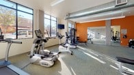 This contemporary fitness center is thoughtfully designed to support a full range of workout routines. The space is anchored by a bright, open cardio area lined with large windows that flood the floor with natural light and offer views of the surrounding outdoors. Exposed industrial-style ductwork runs along the ceiling, adding a modern aesthetic contrast to the clean, neutral tones of the concrete-textured walls and patterned carpet flooring. A bold orange accent wall energizes the strength-training zone, which is equipped with Precor and TRUE brand machines, a multi-station cable system, a dumbbell rack, and a variety of medicine balls for functional training. Adjustable benches provide versatility for both free weight and machine-based exercises. A wall-mounted flat-screen television keeps users entertained during cardio sessions. A large mirrored studio or flex area adjoins the main floor, expanding the usable workout space and offering an ideal environment for stretching, yoga, or personal training. The overall ambiance is clean, energizing, and well-lit, combining industrial design elements with practical fitness amenities to create a motivating environment for users of all fitness levels.