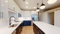 Located at 600 Willard St in Durham, NC, this contemporary two-story home combines modern design with practical functionality throughout its well-appointed layout. The heart of the home is the striking kitchen, which features a two-tone cabinet design pairing crisp white upper cabinetry with deep navy and dark wood lower cabinets. White quartz countertops, a white subway tile backsplash, a matte black pull-down faucet, and a full suite of stainless steel appliances—including a French door refrigerator, range, microwave, and dishwasher—complete the space. A large center island with pendant lighting provides additional prep and gathering space, while light hardwood-style flooring ties the kitchen seamlessly into the open living area. The first floor also includes a half bath with a clean white vanity, black-framed mirror, and matte black fixtures. Upstairs, carpeted bedrooms offer a comfortable retreat, with one room featuring a walk-in closet with wire shelving alongside a dedicated en-suite bath. A stacked washer and dryer unit is conveniently located on the upper level. Additional hallway details include louvered and barn-style doors, adding architectural character throughout the home.