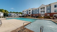 Located at 21 E Revere Blvd in Huntsville, Alabama, this multi-story residential community presents a compelling blend of modern architectural design and thoughtfully curated outdoor amenities. The building exterior showcases a contemporary combination of blue-gray siding, white trim, and brick accents at the base, giving it a polished, upscale appearance. Private balconies extend from multiple levels, offering residents outdoor space with views of the surrounding community and natural treeline.

The centerpiece of the outdoor amenity area is a generously sized resort-style swimming pool with crystal-clear water, framed by a brick-paver pool deck. Ample lounge chairs are arranged throughout the expansive sundeck, providing plenty of space for relaxation under the Alabama sun. A covered pergola structure offers shaded seating for residents seeking a cooler outdoor retreat, complemented by additional patio furniture for casual gatherings.

Manicured landscaping with ornamental shrubs and seasonal plantings borders the outdoor spaces, enhancing the community's curb appeal. Wrought iron fencing encloses the amenity area, providing a sense of privacy and security. The surrounding neighborhood features additional residential buildings and a backdrop of mature trees, contributing to a serene and well-established setting. Overall, this community delivers a refined outdoor living experience in a vibrant Huntsville location.