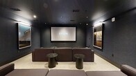 Located at 1000 Spring St NW in Atlanta, GA, this dedicated media room delivers an immersive, cinema-quality experience within a thoughtfully designed space. The room is enveloped in deep charcoal and near-black wall and ceiling tones, creating an intimate, light-controlled environment ideal for screening and entertainment. A large wall-mounted projection screen anchors the front of the room, paired with a ceiling-mounted Epson projector to deliver a true theater experience. Seating is arranged in a tiered, theater-inspired layout comprising generously sized modular gray sectional sofas at the front and a warm cognac leather banquette along the rear wall, accommodating multiple viewers comfortably. Compact round side tables in dark marble and matte black finishes are distributed throughout for added convenience. Recessed ceiling lighting provides subtle ambient illumination, while a series of large, framed photographic art pieces line the walls, adding a curated, gallery-like quality to the space. Light-toned carpeting contrasts the dark walls, softening the overall aesthetic. A sliding door connects the room to an adjacent corridor, ensuring easy access while maintaining the room's enclosed, focused atmosphere.