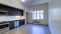 This thoughtfully designed single-floor studio offers a clean, contemporary aesthetic that maximizes every inch of its open-plan layout. The kitchenette is outfitted with dark espresso cabinetry, under-cabinet lighting, a stainless steel sink, and a dishwasher, creating a polished and functional cooking area. Light-toned hardwood-style flooring runs throughout the space, lending warmth and a sense of continuity. A generously sized window allows natural light to flood the interior while framing views of the surrounding urban landscape, including the façade of a neighboring building and greenery below. A wall-mounted PTAC heating and cooling unit sits beneath the window, ensuring year-round climate comfort. The walls are finished in crisp white, complementing the high ceilings that contribute to an airy, open ambiance. A private doorway leads to an adjacent area, likely a bathroom or closet, maintaining a practical flow within the unit. The flush-mount ceiling fixture provides clean overhead lighting, reinforcing the space's modern sensibility. Overall, the studio presents a well-appointed, efficient living environment suited for urban living.
