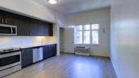 This thoughtfully designed single-floor studio offers a clean, contemporary aesthetic that maximizes every inch of its open-plan layout. The kitchenette is outfitted with dark espresso cabinetry, under-cabinet lighting, a stainless steel sink, and a dishwasher, creating a polished and functional cooking area. Light-toned hardwood-style flooring runs throughout the space, lending warmth and a sense of continuity. A generously sized window allows natural light to flood the interior while framing views of the surrounding urban landscape, including the façade of a neighboring building and greenery below. A wall-mounted PTAC heating and cooling unit sits beneath the window, ensuring year-round climate comfort. The walls are finished in crisp white, complementing the high ceilings that contribute to an airy, open ambiance. A private doorway leads to an adjacent area, likely a bathroom or closet, maintaining a practical flow within the unit. The flush-mount ceiling fixture provides clean overhead lighting, reinforcing the space's modern sensibility. Overall, the studio presents a well-appointed, efficient living environment suited for urban living.
