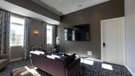 Situated at 2980 Mark Andrew Dr in Dublin, Ohio, this single-floor space makes a strong impression with its thoughtfully designed interior. The lounge area serves as a central gathering space, showcasing deep charcoal and taupe wall tones that create a sophisticated, moody ambiance. A large wall-mounted flat-screen television anchors the main wall, making it an ideal setting for entertainment and relaxation. Plush seating, including a rich plum-toned sofa and additional accent chairs, invites comfortable lounging. Patterned drapery in a bold botanical print frames the windows, allowing natural light to filter softly into the room while adding a decorative focal point. Contemporary wall sconces provide warm accent lighting, complementing the overall refined aesthetic. The flooring features a richly patterned carpet that ties together the color palette throughout the space. Recessed ceiling speakers suggest an integrated audio experience, further enhancing the entertainment-ready atmosphere. Clean white trim and architectural ceiling details add a crisp contrast to the darker wall finishes, lending the space a polished, well-curated character. Overall, the design balances comfort and style in a cohesive, welcoming environment.