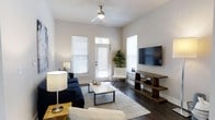 Located at 105 Ivy Green Way in Charleston, South Carolina, this single-story home offers a clean, modern aesthetic with thoughtfully designed living spaces. The living room serves as the heart of the home, showcasing rich dark hardwood flooring that runs throughout and creates a warm, grounding contrast to the crisp white walls. Elevated ceilings add a sense of openness and airiness, while a ceiling fan ensures year-round comfort. Multiple windows dressed with classic white blinds flood the space with natural light, creating a bright and welcoming atmosphere. A glass-paneled exterior door provides direct access to the outdoors, further enhancing the connection between interior and exterior. The living area is well-proportioned, offering ample room for seating arrangements and entertaining. Neutral tones throughout the interior provide a versatile backdrop that suits a variety of design preferences. The overall ambiance is contemporary yet comfortable, reflecting the relaxed charm characteristic of Charleston living. This home combines practical single-floor convenience with stylish finishes, making it an inviting and functional residence in a well-established community.