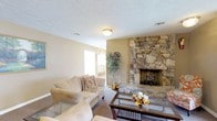 This single-floor residence offers a warm and inviting atmosphere anchored by a standout floor-to-ceiling natural stone fireplace that serves as the living room's dramatic focal point. The fireplace's rugged stonework adds a rustic, timeless character to the space, complementing the neutral beige walls and carpeted flooring throughout. The living area is generously sized, accommodating comfortable seating arrangements with plush sofas and a glass-top coffee table, creating an ideal space for relaxing or entertaining. A textured popcorn ceiling and flush-mount light fixture provide a classic finish overhead, while ceiling vents suggest a well-integrated HVAC system for year-round comfort. Natural light filters into the space through a window, adding brightness to the warm, earthy color palette. A decorative potted plant near the transition to an adjoining area introduces a touch of greenery, enhancing the overall ambiance. The layout flows naturally between areas, offering a cohesive and practical living environment. The combination of stone, soft textiles, and neutral tones gives this home a grounded, comfortable character well-suited for everyday living.