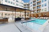 This expansive courtyard amenity space is centrally positioned within a modern multi-story building, combining contemporary design with thoughtfully curated outdoor living areas. The architecture blends white paneling with warm exposed brick facades, creating a visually dynamic backdrop that frames the entire courtyard.

At the heart of the space is a resort-style swimming pool with clear blue water, accessible via stainless steel handrails and surrounded by a broad concrete pool deck lined with lounge chairs for sunbathing. Adjacent to the pool, a covered pergola structure features slatted wood ceilings and ceiling fans, providing shaded seating areas with upholstered chaise lounges and woven accent furniture. This covered zone also incorporates a built-in fireplace and a patterned tile bar area, ideal for social gatherings.

A separate sun-drenched terrace offers additional lounge chairs alongside raised brick planter beds with greenery. Decorative hanging egg chairs add a playful, relaxed element to the courtyard. Floor-to-ceiling glass walls on the building's ground level allow interior common areas to visually connect with the outdoor space.

Herringbone-patterned pavers define the covered lounge zones, while clean concrete walkways guide circulation throughout. The courtyard is open to the sky, allowing abundant natural light to filter throughout the day, creating a vibrant and inviting communal environment.