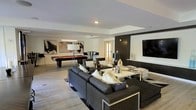 This single-floor space is thoughtfully designed for entertaining, combining multiple functional zones within an open, flowing layout finished in a modern, sophisticated aesthetic. Light hardwood-style flooring runs throughout, unifying the various areas and lending a clean, cohesive feel to the interior.

The central lounge area anchors the space, featuring a large sectional sofa in dark charcoal upholstery with chrome accents, paired with matching armchairs and a set of low-profile coffee and side tables. A large flat-screen television is mounted on a dark accent wall panel, creating a dedicated media focal point. Recessed lighting illuminates the space from above, complemented by strategically placed floor and table lamps.

Adjacent to the lounge, a sleek bar-height dining table with industrial-style stools provides a casual seating option, set beneath large windows dressed with dark curtains that frame the natural light. Bold abstract artwork adorns the walls, adding visual interest throughout.

A dedicated game room area features a full-size pool table with a vibrant red felt surface, completing the entertainment-focused design. The overall ambiance is contemporary and upscale, with a confident, lounge-like atmosphere suited for both relaxation and social gatherings.