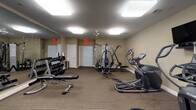 This fitness center offers a comprehensive and well-organized workout environment designed to support a full range of exercise routines. The space is contained within a single, generously sized room finished with neutral beige walls, striped low-pile carpeting, and flush-mounted ceiling light panels that provide consistent, even illumination throughout. Ceiling fans are positioned overhead to maintain comfortable airflow during workouts.

The cardio section features multiple treadmills with individual display consoles, a row of Octane elliptical trainers, and additional low-impact cardio equipment. Each cardio station is paired with a wall-mounted flat-screen television, making it easy to stay entertained during longer sessions. A multi-function cable resistance machine anchors the strength training area, accompanied by a full rack of dumbbells in graduated weights, a flat bench, and a functional trainer for a variety of resistance exercises.

Large wall mirrors along one side of the room visually expand the space and allow users to monitor their form. The room includes clearly marked exit doors and a hand sanitizer station near the entrance, reflecting attention to cleanliness and safety. A window along one wall allows natural light to filter in. Overall, the fitness center presents a clean, functional, and well-maintained environment suitable for a broad range of fitness levels and training goals.