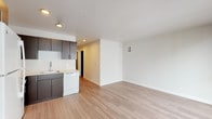 This single-floor unit offers a clean, modern aesthetic defined by light wood-style flooring and crisp white walls that run throughout the space. The open-plan layout seamlessly connects the kitchen and living area, creating a sense of openness and versatility. The kitchen is well-equipped, featuring dark espresso cabinetry with sleek hardware, light countertops, and a full suite of white appliances including a refrigerator, range, microwave, dishwasher, and sink. Recessed lighting illuminates the kitchen and adjoining areas, adding a polished finish. The living area is generously proportioned and bathed in natural light, courtesy of large floor-to-ceiling windows that look out toward the surrounding building exterior. The bright, neutral palette of the living space provides a welcoming and adaptable environment. A hallway connects the main living areas to additional rooms, including a bathroom that continues the contemporary design language with dark wood vanity cabinetry, a white countertop with an integrated sink, and wood-style flooring. The overall ambiance of this unit is modern, clean, and move-in ready, with a cohesive design that balances functionality and style.