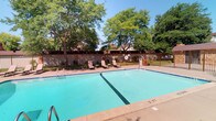 This single-level facility offers a thoughtfully designed combination of outdoor recreation and functional interior amenities. The centerpiece of the outdoor area is a generously sized swimming pool with clear, blue water, flanked by a spacious concrete pool deck. Lounge chairs are positioned poolside, inviting relaxation beneath the shade of mature, leafy trees that frame the space and provide natural privacy. A wrought-iron perimeter fence encloses the area, adding a sense of security while maintaining an open, airy atmosphere under clear skies.

Inside, the restroom facilities reflect a modern, professional design aesthetic. Bold black subway tiles line the lower half of the walls, creating a striking contrast against the crisp white upper walls. Stainless steel fixtures and hardware — including partitioned stalls, urinals, paper towel dispensers, and waste bins — lend a clean, utilitarian elegance throughout. The stall partitions are constructed of brushed stainless steel panels, contributing to the cohesive monochromatic palette. Light-toned tile flooring completes the look, while framed mirrors and a white sink add practical functionality. The overall design balances durability with contemporary style, making this a polished and well-maintained shared facility.