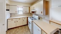 This single-floor unit offers a practical and well-organized layout designed for comfortable everyday living. The kitchen is equipped with white cabinetry accented by warm wood-tone handles and trim, providing generous storage across both upper and lower cabinets. A white gas range with overhead ventilation hood serves as the focal point, complemented by continuous light-colored countertops and a window fitted with blinds that brings in natural light. The flooring features neutral tile-pattern vinyl, adding durability to the space.

The living area is carpeted in a warm beige tone and benefits from direct outdoor access through a glass-paneled exterior door, alongside an adjacent window that fills the room with natural light and views of the surroundings. A second carpeted room, suitable as a bedroom, includes a windowed wall and a closet with bifold doors. Textured ceilings are present throughout the living and sleeping areas.

A dedicated area near the living space features a light wood half-wall or bar partition, with in-unit washer and dryer connections visible beyond. The bathroom includes tile-pattern flooring, a window for ventilation, and an electrical panel conveniently located nearby. Overall, the unit presents a clean, neutral palette throughout.
