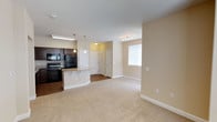 This single-floor residence is designed with an open-concept layout that seamlessly connects the living and kitchen areas. The kitchen is well-appointed with rich, dark-stained cabinetry offering generous storage, granite countertops, and a full suite of black appliances including a gas range, built-in microwave, and refrigerator. A peninsula with a granite surface and pendant lighting above serves as both a functional workspace and an informal dining area. The flooring in the kitchen transitions from hardwood-style planks to plush neutral carpeting in the living area, adding warmth and comfort underfoot.

The living space is open and airy, with neutral beige walls and track lighting that complement the natural light filtering in through a window fitted with blinds. The layout provides a clear sightline from the living area through the kitchen, reinforcing the spacious, connected feel of the unit.

The bedrooms are carpeted and finished with the same warm neutral palette. Multiple windows allow natural light to brighten each room, enhancing the overall sense of space. The bathroom includes a full soaking tub and shower combination with clean white tile surround and built-in shelving, maintaining a simple, functional aesthetic throughout.
