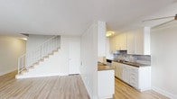 This two-story residence is designed with a clean, functional layout spread across two levels. The main floor features an open living area with light-toned hardwood-style flooring that extends throughout, creating a cohesive and airy atmosphere. A staircase with a white railing connects the levels, positioned alongside a closet door for added storage convenience. The kitchen is well-appointed with white shaker-style cabinetry, dark countertops, a tile backsplash, stainless steel appliances including a refrigerator and dishwasher, and a double basin sink. A peninsula counter extends into the living space, providing a natural division between the kitchen and main living area.

The upper level hosts multiple bedrooms, each carpeted in neutral tones for comfort. The bedrooms feature mirrored sliding closet doors offering generous storage, and windows that allow natural light to filter in. One bedroom includes access to an adjacent room, potentially a secondary space or walk-in area. The bathroom on this level is finished with a white vanity, wood-look flooring, a tub-shower combination with tiled surround, and a small window for ventilation. Throughout, the space maintains a neutral, move-in-ready aesthetic.