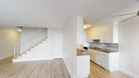 This two-story residence is designed with a clean, functional layout spread across two levels. The main floor features an open living area with light-toned hardwood-style flooring that extends throughout, creating a cohesive and airy atmosphere. A staircase with a white railing connects the levels, positioned alongside a closet door for added storage convenience. The kitchen is well-appointed with white shaker-style cabinetry, dark countertops, a tile backsplash, stainless steel appliances including a refrigerator and dishwasher, and a double basin sink. A peninsula counter extends into the living space, providing a natural division between the kitchen and main living area.

The upper level hosts multiple bedrooms, each carpeted in neutral tones for comfort. The bedrooms feature mirrored sliding closet doors offering generous storage, and windows that allow natural light to filter in. One bedroom includes access to an adjacent room, potentially a secondary space or walk-in area. The bathroom on this level is finished with a white vanity, wood-look flooring, a tub-shower combination with tiled surround, and a small window for ventilation. Throughout, the space maintains a neutral, move-in-ready aesthetic.
