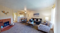 This single-floor residence offers a well-organized and cohesive layout with a warm, coastal-inspired aesthetic carried throughout each room. The living area is generously sized, featuring plush carpeting, a textured ceiling, and a ceiling fan that adds both function and character. Natural light filters in through large vertical-blind-covered windows and a sliding glass door, lending the space an open, airy feel. A standout feature of the living area is a tile-surround fireplace with classic terracotta-toned tiles, providing a cozy focal point. The dining area sits adjacent to the living space, offering a seamless flow ideal for everyday living and entertaining. A pass-through counter connects the dining area to the galley-style kitchen, which is efficiently designed with warm wood cabinetry, white appliances, ample countertop workspace, and durable hardwood-style flooring. The bedroom is comfortably proportioned with soft carpet, a large window that invites natural light, and a relaxed, restful ambiance. The bathroom features a wide vanity with wood cabinetry, a generous countertop, and a large mirror with bright overhead lighting. Overall, the space maintains a consistent, inviting character with neutral tones and practical finishes throughout.