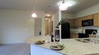 This single-floor residence offers a well-designed open-concept layout that seamlessly connects the kitchen and living areas, creating a spacious and inviting atmosphere. The kitchen is a standout feature, showcasing granite countertops with subtle speckled detailing that adds both elegance and durability to the space. A large kitchen island serves as the centerpiece, ideal for casual dining or entertaining, and is illuminated by stylish pendant lights that cast a warm, focused glow. Additional overhead lighting ensures the space remains bright and functional throughout. The kitchen is equipped with medium-toned wood cabinetry offering ample storage, and a black refrigerator anchors the cooking area with a bold contrast against the light walls. Neutral, warm-toned walls and flooring run throughout the space, contributing to a cohesive and calming ambiance. A hallway visible from the kitchen leads to additional rooms, hinting at a thoughtful and practical floor plan. The overall design balances comfort with functionality, making it a welcoming space suited for both everyday living and entertaining guests.