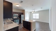 Located at 1680 Hero Way in Leander, Texas, this single-story home offers a well-designed, open-concept layout that seamlessly connects the kitchen, dining, and living areas. The kitchen is a standout feature, outfitted with rich dark cabinetry, granite countertops, a tile backsplash, and a full suite of stainless steel appliances including a range, microwave, and refrigerator. A central kitchen island provides additional counter space and storage, making it highly functional for both cooking and entertaining. Pendant lighting above the island adds a modern, polished touch. The open living area is bright and welcoming, benefiting from natural light through white-trimmed windows and a ceiling fan that enhances comfort. Wood-look plank flooring runs throughout the main living spaces, offering a warm aesthetic that is also easy to maintain. The bathroom features a well-lit vanity with ample counter space. The floor plan's open design ensures a smooth flow between rooms, creating an inviting and practical living environment. The home's clean, neutral color palette and contemporary finishes make it a versatile and move-in ready space.