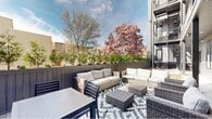 This outdoor terrace is a thoughtfully designed communal space set against the backdrop of a modern multi-story building. The terrace is divided into two distinct functional zones: a furnished lounge and seating area, and a green roof section with low-growing ground cover plants that add a naturalistic counterpoint to the surrounding hardscape.

The lounge area is anchored by a generous arrangement of wicker sectional sofas, armchairs, and a low coffee table, all dressed in neutral cream cushions. A bold black-and-white geometric patterned rug defines the seating area atop a light grey composite deck, while a compact bistro-style dining table and chairs provide an additional gathering spot. The perimeter is enclosed by dark vertical board fencing, lined with raised planters filled with low shrubs and greenery, creating a sense of privacy while framing views of striking blooming magnolia trees beyond.

The covered section of the terrace, tucked beneath an upper-level balcony, features a dark slatted ceiling with recessed and track lighting, extending the usability of the space into the evening. The modern exterior cladding in grey horizontal siding, dark metal accents, and glazed entry doors complete the contemporary aesthetic, resulting in a well-balanced outdoor environment suited for relaxation and social gatherings.