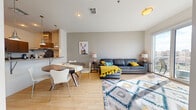 This contemporary single-floor residence offers a well-designed open-concept layout that seamlessly connects the living, dining, and kitchen areas. Light hardwood flooring runs throughout the main living space, enhancing the bright and airy atmosphere created by abundant natural light. The living area is generously proportioned, anchored by large sliding glass doors that open onto a private balcony overlooking an urban cityscape.

The kitchen is thoughtfully appointed with rich wood cabinetry, granite countertops, stainless steel appliances including a range, dishwasher, refrigerator, and microwave, and stylish pendant lighting above a peninsula that provides additional counter space and casual seating. An adjacent dining area comfortably accommodates a round table and chairs.

The residence includes two bedrooms, each featuring plush carpet flooring, ceiling fans, and windows that allow in natural light and urban views. The bathroom is finished with large-format tile, a full soaking tub and shower combination, granite vanity countertop, and access to a walk-in closet. A bold blue entry door adds a distinctive design touch at the front entrance. Overall, the space presents a clean, modern aesthetic with a warm and inviting ambiance throughout.