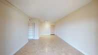 This single-floor space presents a clean and functional layout with a bright, open feel throughout. The main living area is finished with large, light-toned ceramic tiles that extend across the floor, contributing to a sense of spaciousness and easy maintenance. Neutral cream-colored walls and white trim provide a versatile backdrop that suits a wide range of interior styles. A track lighting fixture mounted to the ceiling offers practical, directional illumination, while the smooth white ceiling enhances the room's overall brightness. A notable built-in shelving unit with multiple tiers is accessible through a bifold door, providing convenient and organized storage directly within the living space. The entry door is centrally positioned along the far wall, contributing to a straightforward and logical flow through the unit. Baseboards and subtle architectural lines maintain a tidy, well-kept appearance throughout. The overall ambiance is light, airy, and move-in ready, making it well-suited for those seeking a low-maintenance space with practical features and a neutral palette that allows for flexible furnishing and décor choices.