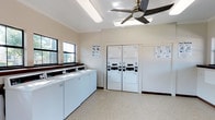 This shared laundry facility offers a clean, well-organized space designed for convenient, efficient use. A row of top-load washing machines lines one wall, while stacked dryer units are positioned along the opposite side, providing ample capacity for residents. The room features a neutral, light-toned color scheme with speckled flooring that contributes to a tidy, functional atmosphere. A ceiling fan with an integrated light fixture helps maintain air circulation throughout the space. Natural light enters through multiple windows along one wall, brightening the interior and creating a comfortable environment. Informational signage is posted near the machines to guide users through proper operation and drying instructions. The layout is open and spacious, allowing easy movement between machines. The facility appears well-maintained, with equipment from a recognized commercial laundry service provider, ensuring reliable performance for day-to-day use.