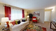 This single-floor residence offers a thoughtfully designed layout that balances comfort and personality across its key living spaces. The living room serves as the heart of the home, featuring plush carpeted flooring, large windows dressed with bold crimson curtains that invite generous natural light, and a textured ceiling. A white sofa anchored by a vibrant patterned area rug, a striking red accent chair, and colorful wall art create a lively yet inviting atmosphere. Adjacent to the living room, the dining area makes a bold statement with a warm burnt-orange accent wall, hardwood flooring, and a round glass dining table surrounded by green upholstered chairs — an ideal space for intimate gatherings. The kitchen is efficiently appointed with white cabinetry, granite-look countertops, and a white appliance suite, and opens directly to the dining area for seamless flow. The bedroom is a serene retreat featuring a ceiling fan, carpeted flooring, patterned drapery, and a rich gold accent wall complemented by dark furnishings and layered artwork. The bathroom offers a full tub-shower combination with ample cabinetry and hardwood-style flooring. Throughout the residence, eclectic art and bold color choices lend a distinctive, curated character.