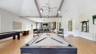 This single-floor amenity space is designed for both recreation and productivity, combining modern aesthetics with a welcoming, social atmosphere. The centerpiece of the main game room is a full-size pool table positioned beneath a striking contemporary pendant light fixture, complemented by a shuffleboard table and an arcade-style game, making it an ideal hub for entertainment. Weathered gray exposed wood beams accent the soaring vaulted ceilings, adding architectural character and a sense of openness throughout the space. Light wood-tone flooring runs seamlessly through all areas, lending warmth and cohesion to the overall design. Abundant natural light pours in through multiple large windows fitted with white blinds, with select windows offering views of outdoor greenery and access to an exterior balcony or patio. A lounge and dining area with high-top tables and mixed seating provides a casual gathering spot. The built-in kitchenette features dark granite countertops, a stainless steel sink, and integrated appliances, making the space well-equipped for hosting. Curated contemporary artwork adorns the walls throughout, enhancing the polished, modern aesthetic. A separate, quietly positioned workstation room offers two computer setups beside a large window, providing a bright and focused environment for productivity within the same convenient facility.