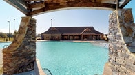 This community amenity center is a standout outdoor facility designed to serve as a central gathering hub. At the heart of the space is a generously sized swimming pool with clear turquoise water, framed by a wide concrete pool deck that provides ample room for lounging and recreation. The pool transitions from shallow to deeper water, as indicated by depth markers along the edge, making it suitable for a range of users.

Defining architectural features include striking stacked-stone pergola columns that support rustic timber archways at the pool's entrance. These natural stone pillars add a refined, lodge-inspired aesthetic that carries throughout the property. The pergola structure frames panoramic views of the pool and surrounding landscape, creating a dramatic focal point upon arrival.

The craftsman-style clubhouse anchors the far end of the pool area, featuring a steeply pitched multi-gabled roofline, exposed timber accents, and large windows that open the interior to natural light. A covered patio runs along the clubhouse's pool-facing side, offering shaded seating and direct access to the deck.

The surrounding grounds are enclosed by black iron fencing and feature open green space with mature trees visible in the distance. Sports court lighting poles are positioned throughout the exterior, extending the usability of the facility into evening hours. The overall ambiance is open, welcoming, and thoughtfully designed for community enjoyment.