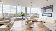 This sophisticated upper-floor amenity space offers an expansive, open-concept layout designed for both relaxation and social gathering. Abundant natural light pours in through dramatic floor-to-ceiling windows that wrap around the space, framing panoramic views of the surrounding cityscape and autumn treetops beyond. Light oak hardwood flooring runs throughout, complementing the crisp white walls and clean architectural lines.

The lounge area is anchored by a generously sized sectional sofa and coordinating armchairs in neutral tones, arranged around a glass-and-metal coffee table, with a sleek wall-mounted fireplace set against a marble surround adding a refined focal point. A long hallway lined with curated artwork connects the lounge to the rest of the floor, maintaining a gallery-like aesthetic throughout.

The kitchen and dining area features a bold navy cabinetry palette contrasted by white subway tile backsplash, a large island with teal bar seating, and multiple pendant light fixtures. Several dining tables with contemporary grey and teal chairs provide ample seating for group gatherings.

Large windows in every zone offer direct sightlines to the outdoor terrace, which is furnished with patio seating and accented by vibrant red umbrellas. The overall ambiance is bright, airy, and modern — a polished communal environment designed for elevated everyday living.