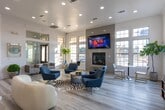 Located at 30000 FM-2978 in Magnolia, Texas, this single-story community clubhouse is designed to deliver a welcoming and functional gathering space with upscale finishes. The open-concept layout is anchored by light wood-look flooring, crisp white walls, and an abundance of natural light streaming through large multi-pane windows that frame views of the surrounding landscape.

The main lounge area is a standout feature, boasting soaring ceilings, recessed lighting, and a sleek fireplace set beneath a wall-mounted display screen. Stylish navy blue accent chairs, bold artwork, and eclectic décor create an inviting atmosphere suited for both relaxation and socializing. A separate seating nook with floor-to-ceiling windows and gold-accented bistro furniture adds an airy, café-inspired touch.

The kitchen is well-appointed with raised-panel white cabinetry, white quartz countertops, a stainless steel farmhouse sink, and stainless steel appliances including a dishwasher and refrigerator. Colorful pendant lighting adds a playful contrast to the otherwise clean, neutral palette. Arched doorways and decorative columns lend architectural character throughout the open floor plan.

A dedicated workspace features a built-in desk, custom open shelving, natural light from a window, and a warm gold pendant light — ideal for remote work or study. The overall ambiance is polished yet approachable, blending contemporary design with comfortable, community-focused functionality.