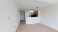 This single-floor space features an open-concept layout that connects the living area to the kitchen, creating a cohesive and functional flow throughout. The living area is finished with neutral-toned wall-to-wall carpeting, contributing to a clean and understated aesthetic. White walls and a white ceiling enhance the sense of openness, while recessed lighting provides overhead illumination.

The kitchen is defined by a peninsula-style pass-through counter with a white exterior, which serves as both a visual divider and a practical surface. Inside the kitchen, warm-toned wood cabinetry lines the upper walls, paired with black appliances including a range and built-in microwave. A stainless steel refrigerator adds a contrasting finish. A fluorescent light fixture is mounted beneath the upper cabinetry, providing direct task lighting over the workspace.

A hallway extends from the living area toward additional rooms, suggesting a practical floor plan with clear circulation paths. A door visible at the end of the hallway provides access to other areas of the unit. Overall, the space presents a straightforward, functional design with a neutral palette suited to a variety of interior styles.