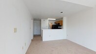 This single-floor space features an open-concept layout that connects the living area to the kitchen, creating a cohesive and functional flow throughout. The living area is finished with neutral-toned wall-to-wall carpeting, contributing to a clean and understated aesthetic. White walls and a white ceiling enhance the sense of openness, while recessed lighting provides overhead illumination.

The kitchen is defined by a peninsula-style pass-through counter with a white exterior, which serves as both a visual divider and a practical surface. Inside the kitchen, warm-toned wood cabinetry lines the upper walls, paired with black appliances including a range and built-in microwave. A stainless steel refrigerator adds a contrasting finish. A fluorescent light fixture is mounted beneath the upper cabinetry, providing direct task lighting over the workspace.

A hallway extends from the living area toward additional rooms, suggesting a practical floor plan with clear circulation paths. A door visible at the end of the hallway provides access to other areas of the unit. Overall, the space presents a straightforward, functional design with a neutral palette suited to a variety of interior styles.