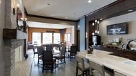 This single-floor hospitality dining space combines contemporary design with a warm, welcoming ambiance. The open layout features a well-defined dining area furnished with round tables and upholstered chairs with dark wood frames, creating an inviting atmosphere for guests. Large windows along the far wall flood the space with natural light, offering views to the outdoors and lending the room an airy, open feel. Warm terracotta accent walls contrast beautifully with cool blue-gray tones, adding visual depth and character throughout. A sleek bar counter anchors one side of the room, lined with high stools and illuminated by elegant pendant lights suspended on cables from the ceiling. Adjacent to the bar, a dedicated beverage station provides guests easy access to coffee and other refreshments. Dark tile flooring runs throughout, grounding the space and complementing the rich, dark wood finishes of the furniture and millwork. Recessed ceiling lighting, combined with the pendant fixtures, creates layered illumination suited to both casual dining and relaxed socializing. Crown molding details add a refined architectural touch. Overall, the space strikes a balance between functional hospitality design and sophisticated, comfortable styling.