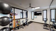 This single-level fitness space is designed to support a well-rounded workout experience. The room features clean, modern finishes with light wood-style flooring, white walls, and dark window trim that lend a contemporary aesthetic throughout.

The space is outfitted with a stationary cycling bike positioned beneath a ceiling fan, which ensures comfortable airflow during intense workouts. A rack of colorful kettlebells and resistance bands provides a variety of strength and conditioning options, while additional equipment visible along the perimeter accommodates diverse training styles.

A large wall-mounted flat-screen television and a smaller interactive display panel are installed on one wall, offering entertainment or guided workout programming. Natural light fills the room through multiple windows fitted with blinds, including a large interior-facing window that overlooks an adjacent space, enhancing the open, connected feel of the facility.

The ceiling fan with an integrated light fixture adds both function and style overhead. The overall layout is open and uncluttered, allowing for ease of movement between equipment. Together, the thoughtful equipment selection, bright lighting, and ventilation create a practical and inviting environment suited for daily fitness routines.