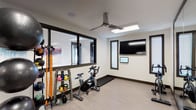 This single-level fitness space is designed to support a well-rounded workout experience. The room features clean, modern finishes with light wood-style flooring, white walls, and dark window trim that lend a contemporary aesthetic throughout.

The space is outfitted with a stationary cycling bike positioned beneath a ceiling fan, which ensures comfortable airflow during intense workouts. A rack of colorful kettlebells and resistance bands provides a variety of strength and conditioning options, while additional equipment visible along the perimeter accommodates diverse training styles.

A large wall-mounted flat-screen television and a smaller interactive display panel are installed on one wall, offering entertainment or guided workout programming. Natural light fills the room through multiple windows fitted with blinds, including a large interior-facing window that overlooks an adjacent space, enhancing the open, connected feel of the facility.

The ceiling fan with an integrated light fixture adds both function and style overhead. The overall layout is open and uncluttered, allowing for ease of movement between equipment. Together, the thoughtful equipment selection, bright lighting, and ventilation create a practical and inviting environment suited for daily fitness routines.