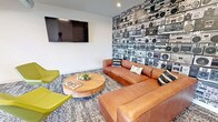Located at 715 Withington Street in Ferndale, Michigan, this single-floor common lounge space presents a thoughtfully designed interior that blends modern aesthetics with bold, expressive detailing. The layout flows across several distinct seating zones, each offering its own character while maintaining a cohesive design language throughout.

A standout feature is the eye-catching accent wall adorned with a floor-to-ceiling boombox-patterned wallcovering, paired with a large wall-mounted television and a generously sized cognac leather sectional sofa atop a layered area rug. A round natural wood coffee table anchors the lounge seating, complemented by chartreuse accent chairs that add a vibrant pop of color.

An adjacent gathering area showcases a sleek linear fireplace set into dark slate tile, flanked by sculptural egg-style lounge chairs and a botanical wallcovering in warm olive tones. Skylight panels and a circular pendant light fixture illuminate the space from above, while the high ceilings lend an open, airy quality.

Near the elevator landing, a expansive floor-to-ceiling window frames panoramic views of the Ferndale skyline and tree canopy below, filling the corridor with abundant natural light. Throughout, patterned carpeting, white walls, and carefully curated furnishings create a welcoming and visually dynamic communal environment.