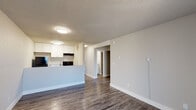This single-floor unit offers a clean, modern layout designed for comfortable everyday living. The open-concept living and kitchen area forms the heart of the space, with durable wood-look flooring running throughout that adds warmth and visual continuity. The kitchen is well-equipped with white upper cabinetry that extends to the ceiling for ample storage, light granite-style countertops, black appliances including a refrigerator and electric range, and a peninsula that separates the kitchen from the living area while providing additional counter space. Flush-mount ceiling lighting keeps the space bright and functional. The living room is generously sized, featuring a wall-mounted air conditioning unit and a sliding glass door dressed with vertical blinds that opens to a private balcony, inviting natural light into the room. The bedroom is a comfortable retreat with a window that brings in additional natural light and a baseboard heating unit for warmth. The bathroom is clean and straightforward, offering a full tub-shower combination with tiled surround and brushed nickel fixtures. Textured ceilings are consistent throughout the unit, and the neutral color palette of warm grays and whites creates a bright, cohesive aesthetic throughout.