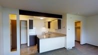 This single-floor space showcases an open-concept kitchen that serves as the focal point of the layout. The kitchen is defined by a peninsula-style counter with a neutral laminate surface, providing both workspace and a natural division between the kitchen and the adjoining living area. Dark espresso-toned cabinetry lines the upper and lower sections of the kitchen, creating a contrast against the white walls and lighter surfaces throughout the space. Integrated appliances, including a range and sink, are neatly built into the countertop run, contributing to a streamlined, functional design. A bold dark structural beam runs across the ceiling above the kitchen, adding an architectural detail that draws the eye and adds visual interest to the otherwise clean, minimal aesthetic. Recessed storage areas are visible on either side of the kitchen, offering organizational utility. The flooring transitions between tile in the kitchen area and carpet in the adjoining living space, delineating different zones within the open plan. Overhead lighting provides steady illumination throughout, and the overall ambiance is practical and uncluttered.