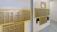 This shared amenity area is situated within a covered parking structure and serves as a central hub for two essential resident services: mail collection and on-site laundry. The space features a straightforward, utilitarian design with smooth, painted concrete walls and durable concrete flooring throughout.

The mail area is the focal point of this space, featuring multiple wall-mounted mailbox units framed in bright yellow accents. The tan-toned metal mailboxes are arranged in tiered rows, each clearly labeled and equipped with individual locks for secure mail retrieval. Outgoing mail slots and a cork bulletin board for community notices are also present, adding to the area's functionality.

Adjacent to the mail area, a dedicated laundry room is accessible through a clearly marked doorway. The laundry room features checkered tile flooring and houses laundry equipment for resident use. A warning sign is posted near the entrance, indicating building-specific regulations for the space.

The surrounding covered parking structure provides overhead protection and is marked with painted yellow lane lines and columns. The overall ambiance is clean and practical, prioritizing resident convenience within a well-organized shared facility.