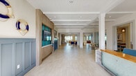 Located at 1250 Marden Rd in Apopka, FL, this generously sized single-floor amenity space offers a sophisticated coastal aesthetic defined by light wood-look tile flooring, crisp white walls, and an abundance of natural light throughout. The grand entry corridor welcomes visitors with warm wood paneling, built-in display screens, and coffered ceilings that set an inviting tone. The centerpiece of the space is a dramatic double-height living room anchored by a floor-to-ceiling stacked stone fireplace with a mounted flat-screen TV. Plush seating arrangements, layered area rugs, and curated coastal artwork create a relaxed yet refined atmosphere. An open-plan kitchen and lounge area features white shaker cabinetry, a blue tile backsplash, quartz countertops, a beverage refrigerator, and a long bar-height peninsula with barstool seating. Adjacent casual seating nooks with tufted banquettes and pedestal tables offer views of palm trees through large windows. A glass-enclosed conference or private dining room provides a flexible, well-lit meeting space. Structural columns and exposed beamed ceiling accents reinforce the architectural character, while an upper-level mezzanine overlooks the main living area, adding vertical drama and openness to the overall design.