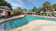 Located at 2181 Dunlap St in North Charleston, South Carolina, this outdoor recreational facility serves as the pool amenity area for Plantation Flats. The centerpiece is a generously sized freeform swimming pool with striking turquoise water, finished with a classic brick coping edge and a wide concrete pool deck that provides ample space for lounging and movement. A row of chaise lounge chairs lines the perimeter, offering residents dedicated sunbathing space. A white pergola structure anchors one side of the deck, adding architectural charm and partial shade to the outdoor area. The pool is flanked by lush mature landscaping, including tall palm trees and dense greenery, which lend a tropical, resort-like ambiance. A single-story brick clubhouse building adjoins the pool area, featuring a glass-paneled entry door and serves as the pool house. Outdoor wicker lounge furniture is positioned near the building's entrance, providing a comfortable gathering area. Dedicated restroom facilities, including a women's restroom, are accessible directly from the pool deck. The overall setting is bright and open, bathed in natural sunlight and framed by the natural beauty of the surrounding tree canopy, creating an inviting and relaxed outdoor retreat.