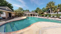 Located at 2181 Dunlap St in North Charleston, South Carolina, this outdoor recreational facility serves as the pool amenity area for Plantation Flats. The centerpiece is a generously sized freeform swimming pool with striking turquoise water, finished with a classic brick coping edge and a wide concrete pool deck that provides ample space for lounging and movement. A row of chaise lounge chairs lines the perimeter, offering residents dedicated sunbathing space. A white pergola structure anchors one side of the deck, adding architectural charm and partial shade to the outdoor area. The pool is flanked by lush mature landscaping, including tall palm trees and dense greenery, which lend a tropical, resort-like ambiance. A single-story brick clubhouse building adjoins the pool area, featuring a glass-paneled entry door and serves as the pool house. Outdoor wicker lounge furniture is positioned near the building's entrance, providing a comfortable gathering area. Dedicated restroom facilities, including a women's restroom, are accessible directly from the pool deck. The overall setting is bright and open, bathed in natural sunlight and framed by the natural beauty of the surrounding tree canopy, creating an inviting and relaxed outdoor retreat.