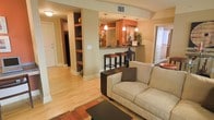 This single-floor residence showcases an open-concept layout that seamlessly connects the living area to the kitchen, creating a cohesive and functional living environment. Light hardwood flooring runs throughout the space, contributing to a bright and inviting ambiance.

The living area features a neutral-toned sofa paired with a dark coffee table and a geometric area rug in warm earth tones, anchoring the space with a comfortable yet stylish aesthetic. A dark side table provides additional surface area beside the sofa.

A structural column with built-in open shelving serves as a subtle divider between the living area and a hallway, offering both practical storage and a decorative display space. The warm beige and terracotta wall tones lend a rich, welcoming character to the interior.

The kitchen is accessible via a raised breakfast bar with dark bar stools, illuminated by pendant lighting hanging from above. The kitchen's terracotta accent wall and additional pendant fixtures add depth and warmth to the cooking space.

A hallway visible from the living area leads to additional rooms, with what appears to be a closet or storage area integrated into the corridor, maximizing the utility of the space.
