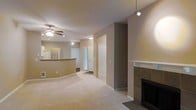 This single-floor residence offers a well-designed open-concept layout that seamlessly connects the main living areas. The spacious living room is anchored by a classic tiled fireplace with a clean white mantle, providing a focal point for the room. Plush wall-to-wall carpeting runs throughout the living space, adding warmth and comfort underfoot.

The kitchen is positioned adjacent to the living area and features a curved breakfast bar that serves as a natural divider between the two spaces while maintaining an open feel. A refrigerator is visible within the kitchen, and the layout appears functional and efficient.

Overhead, a ceiling fan with an integrated light fixture ensures year-round comfort in the main living area, while a modern pendant track light adds a contemporary touch near the entryway. Natural light filters in through a window near the front door, brightening the space.

The neutral, warm beige tones on the walls and ceilings create a calm, cohesive aesthetic throughout. Multiple doorways lead to additional rooms and closet storage, suggesting a practical floor plan. The front entry area is clearly defined, offering a welcoming transition into the home. Overall, the space presents a clean, move-in-ready environment with a comfortable and functional design.