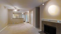 This single-floor residence offers a well-designed open-concept layout that seamlessly connects the main living areas. The spacious living room is anchored by a classic tiled fireplace with a clean white mantle, providing a focal point for the room. Plush wall-to-wall carpeting runs throughout the living space, adding warmth and comfort underfoot.

The kitchen is positioned adjacent to the living area and features a curved breakfast bar that serves as a natural divider between the two spaces while maintaining an open feel. A refrigerator is visible within the kitchen, and the layout appears functional and efficient.

Overhead, a ceiling fan with an integrated light fixture ensures year-round comfort in the main living area, while a modern pendant track light adds a contemporary touch near the entryway. Natural light filters in through a window near the front door, brightening the space.

The neutral, warm beige tones on the walls and ceilings create a calm, cohesive aesthetic throughout. Multiple doorways lead to additional rooms and closet storage, suggesting a practical floor plan. The front entry area is clearly defined, offering a welcoming transition into the home. Overall, the space presents a clean, move-in-ready environment with a comfortable and functional design.