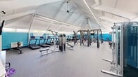 Located at 1 Forest Ridge Rd in Derry, New Hampshire, this fitness facility offers a spacious, single-floor layout designed to accommodate a comprehensive range of workout activities. The space is immediately distinguished by its dramatic vaulted ceiling with exposed white structural beams, which rise to impressive heights and lend the interior an open, airy quality. Clerestory windows lining the upper walls invite abundant natural light, while ceiling fans and modern lighting fixtures ensure comfort throughout the space.

The interior is finished with durable rubber flooring in a neutral gray tone, ideal for high-traffic fitness use. Bold blue accent walls punctuate the perimeter, creating an energizing and motivating environment. A reception or check-in counter is positioned near the entrance, alongside organized storage cubbies and a wall-mounted display screen.

The equipment layout is thoughtfully zoned. A dedicated cardio area houses multiple treadmills and step machines with integrated screens. Strength training is well-represented with a multi-station cable machine, adjustable benches, a full dumbbell rack, and a multi-functional power rack stocked with kettlebells, medicine balls, and resistance accessories. A separate area accommodates group fitness with yoga mats and stability balls. Yellow-trimmed entry doors add a cheerful finishing touch to this clean and well-organized facility.