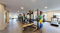 Located in Woodinville, Washington, this well-appointed fitness center offers a generously sized, single-floor exercise space designed to accommodate a wide range of workout needs. The open floor plan is anchored by structural columns finished in warm wood tones, lending the space a modern, clean aesthetic that complements the neutral-toned carpeted flooring throughout.

The cardio zone features a row of elliptical trainers and treadmills positioned along the windowed perimeter, allowing natural light to fill the space and providing views of the surrounding greenery. The strength training area is equipped with multiple cable machines, multi-functional racks loaded with weight plates and medicine balls, adjustable benches, and an extensive dumbbell rack with a full mirror behind it for form monitoring.

A dedicated functional training section offers open floor space for free movement and stretching. Built-in wooden shelving units provide organized storage for accessories such as towels and fitness equipment. Recessed ceiling lighting ensures even, bright illumination throughout the entire facility.

The space also includes a tiled accent column serving as an architectural focal point, glass-panel room dividers, and wall-mounted displays. Large windows along multiple walls draw in natural light and connect the interior to the outdoor environment, creating an inviting and energizing atmosphere for workouts.