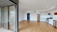 This contemporary single-floor unit showcases a clean, modern design with an open-concept layout that seamlessly connects the living area to the kitchen. Warm-toned hardwood flooring runs throughout, complementing the crisp white walls and recessed lighting overhead. Large floor-to-ceiling glass pivot doors in the living area flood the space with natural light and provide access to an adjacent glazed corridor or balcony, enhancing the sense of openness.

The kitchen is well-appointed with sleek flat-front white cabinetry extending to the ceiling, a full suite of stainless steel appliances including a refrigerator, gas range, built-in microwave, and dishwasher, along with a clean white backsplash and undermount sink.

The bedrooms are bright and airy, with large windows — one offering a view of a palm tree, suggesting a warm-climate setting — and generous closet space. Both bathrooms maintain the unit's modern aesthetic, featuring floating wood-toned vanities with white quartz countertops, undermount sinks, and grey hexagonal tile flooring. One bathroom includes a tub-shower combo with marble-look tile surround, while the second offers a spacious layout with a walk-in shower area.

Overall, the space presents a cohesive, minimalist aesthetic with thoughtful finishes throughout.