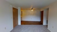 This single-floor residential space offers a straightforward and functional layout designed for comfortable everyday living. The main living area greets you upon entry through a warm wood-toned front door, immediately revealing an open and airy room that combines carpeted and hardwood flooring — a practical blend that defines distinct zones within the space. A ceiling fan with integrated lighting is centrally mounted overhead, providing both air circulation and ambient illumination throughout the area. The neutral, light-colored walls lend the space a clean and versatile backdrop, making it easy to personalize with furnishings and décor. A glimpse into the adjacent kitchen is visible from the living area, suggesting a connected and accessible layout that supports easy movement between spaces. An additional doorway on the far wall leads to further rooms within the unit, hinting at a well-organized floor plan. The overall ambiance is simple and move-in ready, with a calm, understated character well-suited for those seeking a no-frills, functional living environment. The combination of hard and soft flooring surfaces adds a degree of warmth and practicality to the space as a whole.